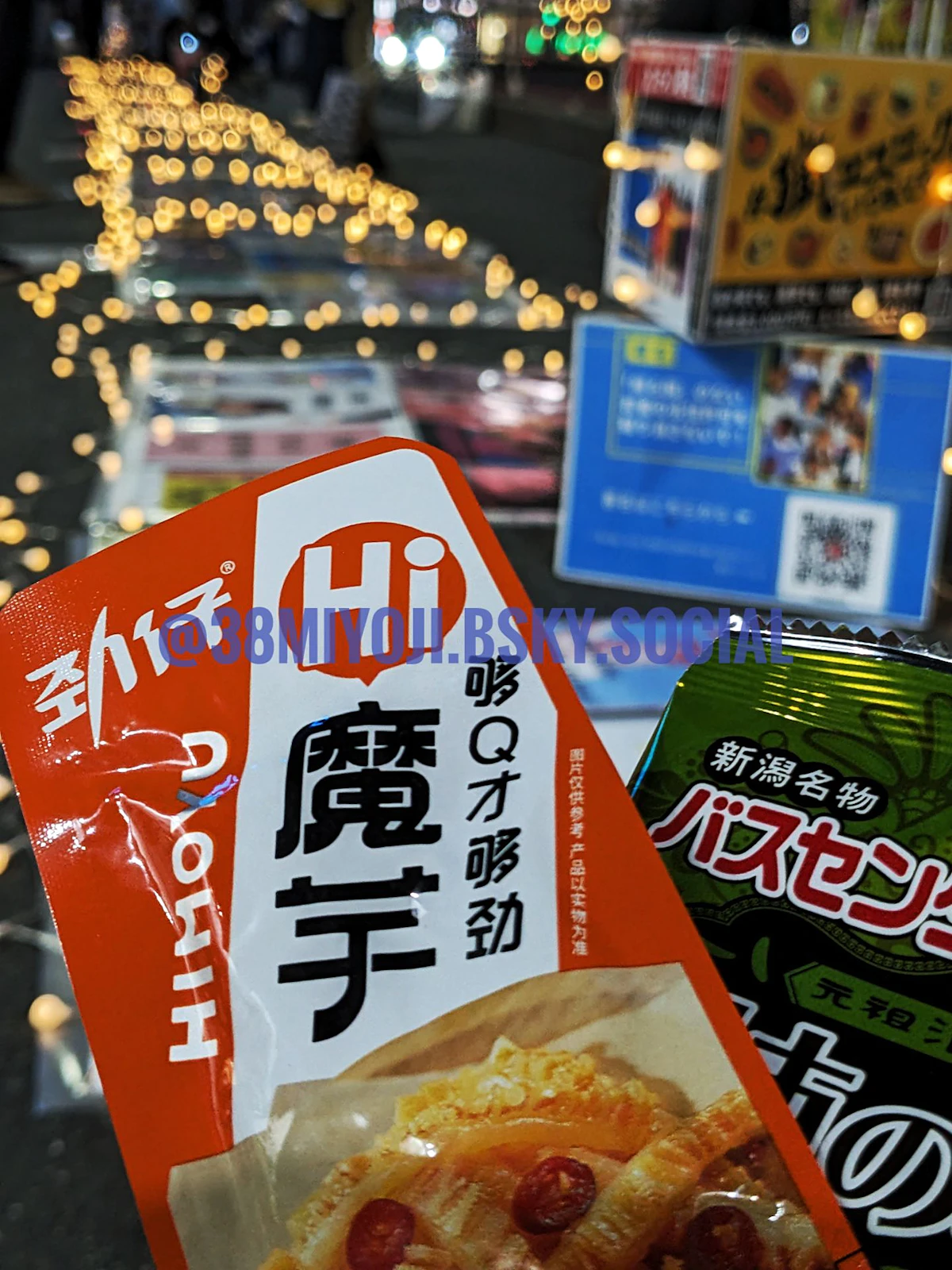 こんにゃくと柿の種のパッケージ2つを手前に、地面に並べられたプラカードの列を奥に捉えた写真。プラカードの周囲にはガーランドライトが張り巡らされている。