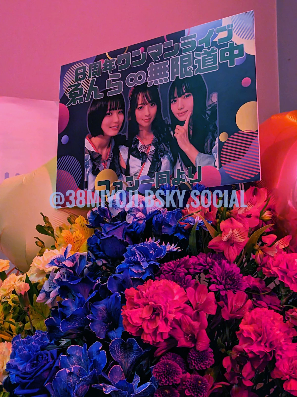 ライブイベントへの祝い花とメッセージボードの写真。赤青黄色の３色の花の上に、メッセージボードが立てられている。メッセージボードの内容は、「８周年ワンマンライブ　ゑんら無限道中　ファン一同より」の文章と、ゑんらメンバー3人の写真。